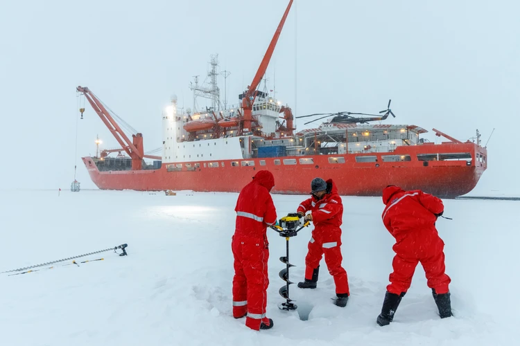 Das Bild zeigt Forschende in roten Schutzanz&uuml;gen, die auf dem Eis Proben entnehmen.